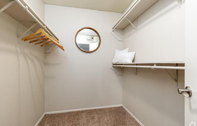 A small, empty closet featuring two shelves, a round mirror on the wall, and a couple of wooden hangers. The floor is covered in light brown carpet, and the walls are painted white. The space is well-lit, providing a clean and minimalist look.