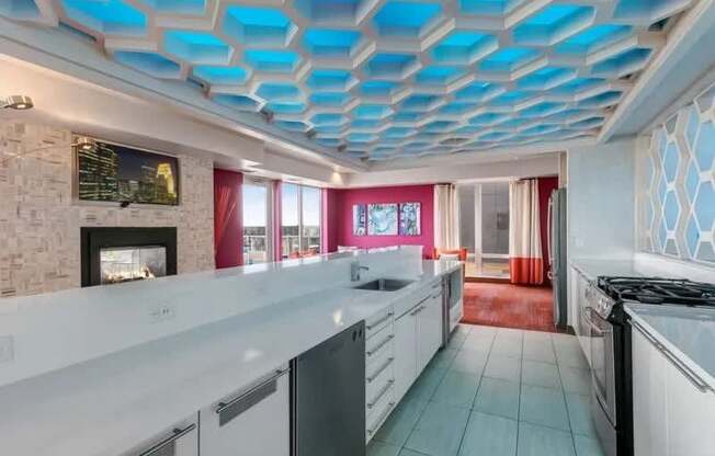 A modern communal kitchen and lounge here at Blue and Lime featuring a long white island with built-in sinks and cabinetry, stainless appliances, and light tile flooring beneath a dramatic blue geometric ceiling. Floor-to-ceiling windows, a fireplace accent wall, and colorful artwork connect the space to a vibrant seating area filled with natural light.