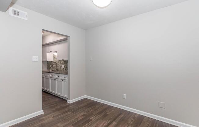 A well-lit room with light gray walls and a wooden floor. The entrance to a kitchen is visible in the background, featuring white cabinets and a tiled backsplash. There is a ceiling light fixture and no furniture in the room, creating an open and spacious feel.