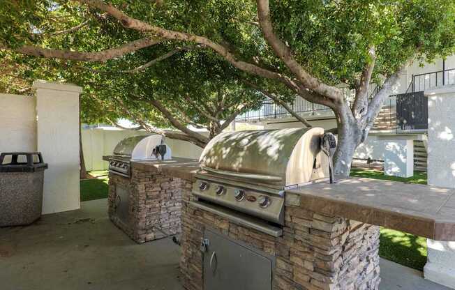 A large outdoor grill is built into a stone wall under a tree.