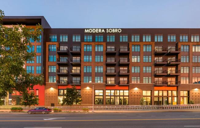 Twilight view of Modera SoBro’s exterior, featuring a bold mix of brick and dark paneling with private balconies and expansive windows.