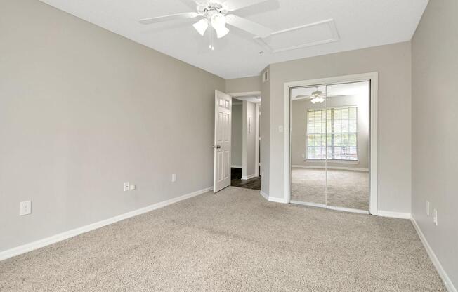 Interior view of a light-colored bedroom with beige carpet. A ceiling fan is visible, and there's a closet with mirrored sliding doors. The room features a neutral wall color and a window with blinds, leading to the adjacent room beyond the doorway. Natural light enhances the space.