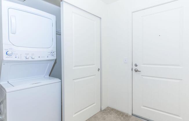A laundry area featuring a stacked washer and dryer unit next to a closed door. The walls are painted white, and there is carpet flooring. A door leading to the outside or another room is visible, with a small peephole. The overall space appears clean and well-maintained.