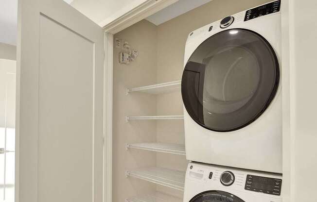 A white washing machine is in a small space between a wall and a shelf.
