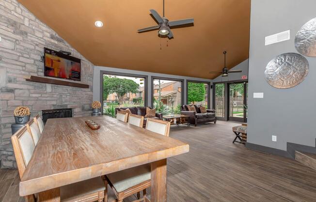 A modern living space featuring a wooden dining table with chairs, a stone fireplace, and a large window view. The ceiling has a warm color and fans, while the decor includes circular wall art and a contemporary TV setup. Lush greenery is visible through the windows, creating an inviting atmosphere.
