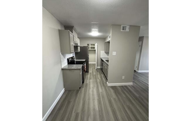 A modern kitchen featuring gray cabinets, stainless steel appliances, and a textured countertop. The open layout includes an area for a refrigerator and a well-lit space. Stylish laminate flooring extends throughout, enhancing the contemporary feel of the design.