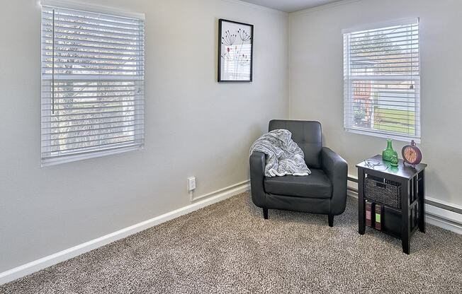 Carpeted living space, with two windows on two walls. Staged with a chair and side table.at Woodhaven, Everett, WA 98203