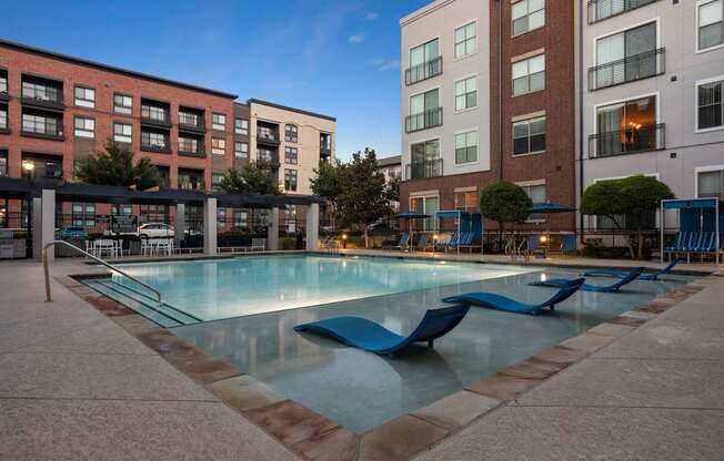 A swimming pool surrounded by a poolside lounge area and apartment buildings.