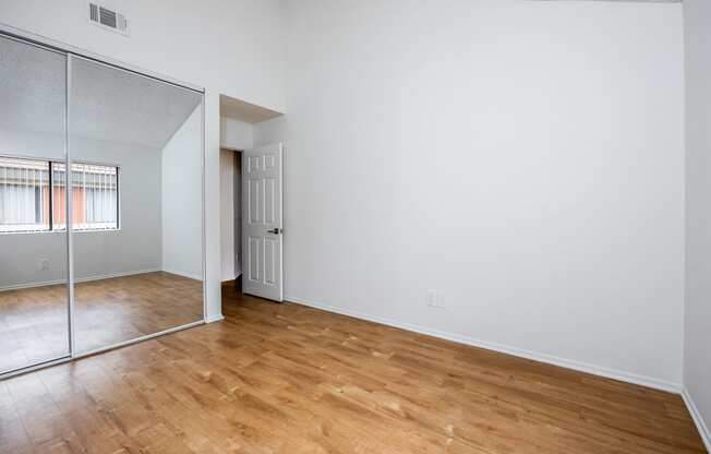 the living room and bedroom of an apartment with wood flooring and a mirrored closet