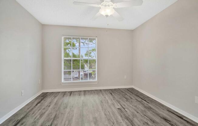 A well-lit empty room featuring light gray walls, a ceiling fan, and a large window with white blinds. The floor is covered with laminate wood flooring. The space appears clean and ready for furnishing.