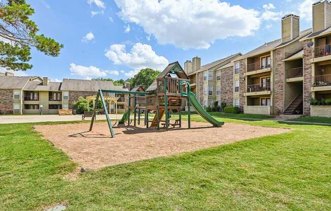 A playground with a green slide and sandbox in front of apartment buildings.