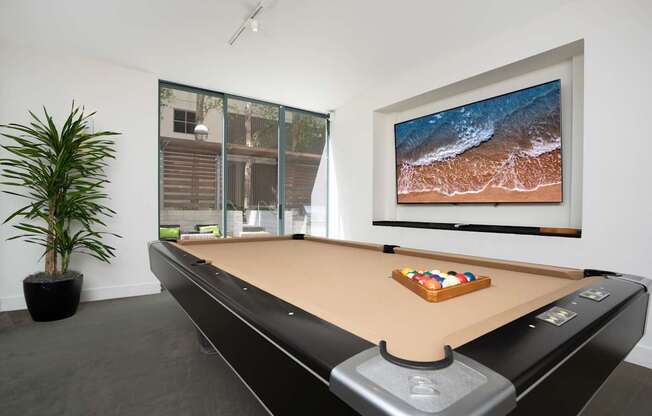 A pool table with a brown cloth and a television on the wall.