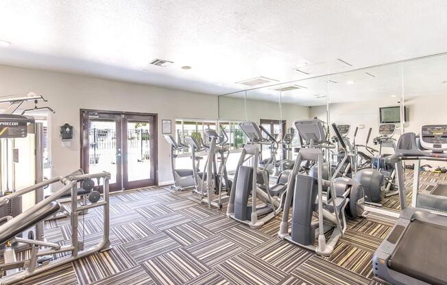 A modern gym interior featuring several exercise machines, including treadmills and elliptical trainers. The space is well-lit with large mirrors on one wall and a patterned floor. Large windows provide views outside, and the environment appears clean and organized, ideal for workout sessions.