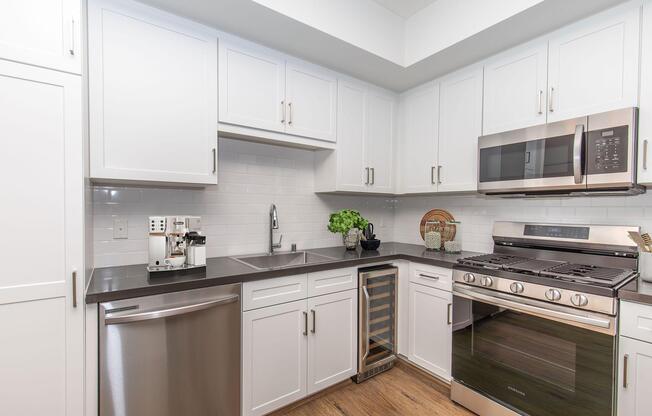 Modern kitchen featuring white cabinets, stainless steel appliances including an oven and refrigerator, a black countertop, a sink, and a wine cooler. The wall is tiled with white subway tiles, and potted plants add a touch of greenery to the space. Hardwood flooring completes the warm, inviting atmosphere.