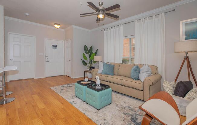 A cozy living room featuring a beige sofa with colorful cushions, a light blue coffee table, and a decorative area rug. There is a lamp and a plant beside the sofa, with a ceiling fan overhead. Natural light filters through the curtains, and a door leads to an entrance area with a small table and chairs.