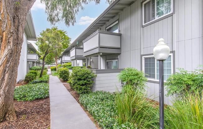 A walkway lined with well-maintained shrubs and grass, leading to modern gray buildings with balconies. Soft landscaping features surrounding the path create a serene atmosphere, complemented by a decorative light post. The scene is bright and inviting, suggesting a pleasant residential area.