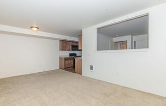 A spacious, empty room with light-colored walls and carpet. There is a kitchen area with wooden cabinets, a stove, and a microwave visible through an opening. The layout is open, and there is a ceiling light fixture. The overall ambiance is clean and ready for furnishing.