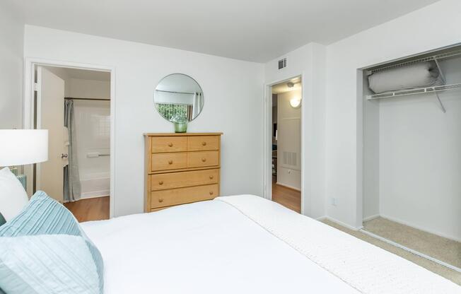 A cozy bedroom featuring a queen-sized bed with light blue pillows, a wooden dresser with five drawers, and a round mirror above it. A doorway leads to a bathroom visible in the background, and another door opens to a spacious closet. The walls are painted white, enhancing the bright, airy atmosphere.