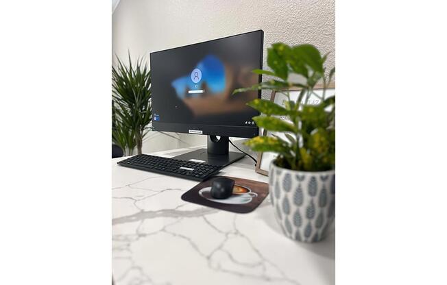 A desk setup featuring a modern computer monitor displaying a login screen, a black keyboard, a mouse on a mousepad, and a small potted plant. The surface is white with a marble pattern, and there are decorative elements in the background, including a tall green plant.