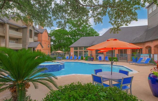A bright and inviting swimming pool surrounded by lush greenery, orange umbrellas, and blue lounge chairs. In the background, there are buildings with a mix of brick and modern architecture. The scene is tranquil and designed for relaxation, with clear blue skies overhead.
