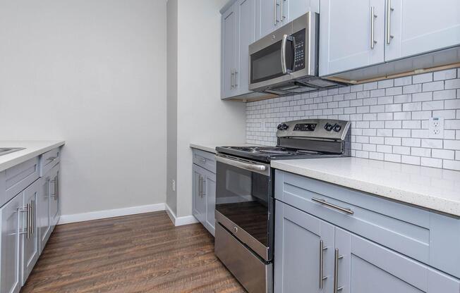 A modern kitchen featuring gray cabinetry, a stainless steel stove and microwave, and a white subway tile backsplash. The countertop is a light-colored quartz, and the floor has a warm wood finish, creating a bright and inviting atmosphere.