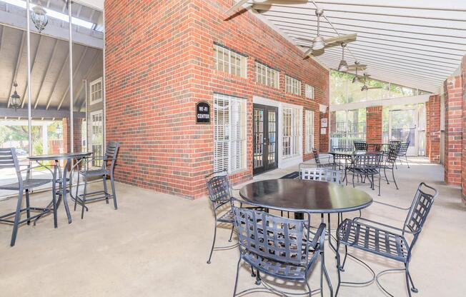 A spacious outdoor seating area featuring black metal tables and chairs, surrounded by red brick walls and large windows. The space is covered by a roof, and there are ceiling fans for ventilation, creating a comfortable atmosphere suitable for gatherings or relaxation.