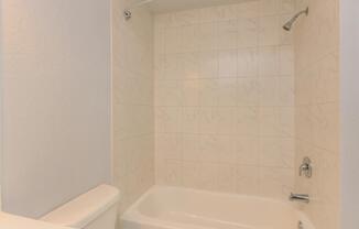 A clean and minimalist bathroom featuring a bathtub with a showerhead above. The walls are covered with light-colored, textured tiles, creating a bright and spacious atmosphere. The floor is unobstructed, emphasizing the simplicity of the design.