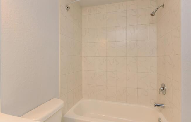 A clean and minimalist bathroom featuring a bathtub with a showerhead above. The walls are covered with light-colored, textured tiles, creating a bright and spacious atmosphere. The floor is unobstructed, emphasizing the simplicity of the design.
