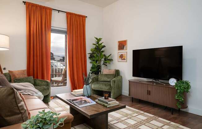 A living room with a brown couch, a green armchair, a coffee table, a flat screen TV, and a window with orange curtains.