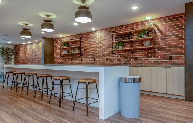 Modern kitchen interior featuring a long white bar with wooden stools, set against a brick wall. Shelves with decorative plants and items are mounted above the bar. The space is well-lit with pendant lights and includes a sleek trash can, creating a stylish and inviting atmosphere.