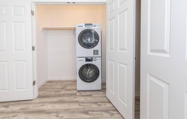 A clean laundry room featuring a stacked washer and dryer, located between open double doors. The room has light-colored walls and a bare wooden shelf above the appliances, set on a light wooden floor.