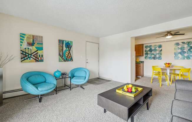 A living room with a grey sofa, a blue chair, a yellow chair, and a table with a bowl of fruit on it.