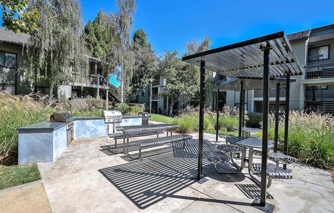 Apartments Hayward For Rent - Outdoor BBQ Area Has Two Gas Grills, Counterspace, Trash Can, Metal Benches, And Shade. The BBQ Area Is Surrounded By Maintained Landscape And Apartment Buildings