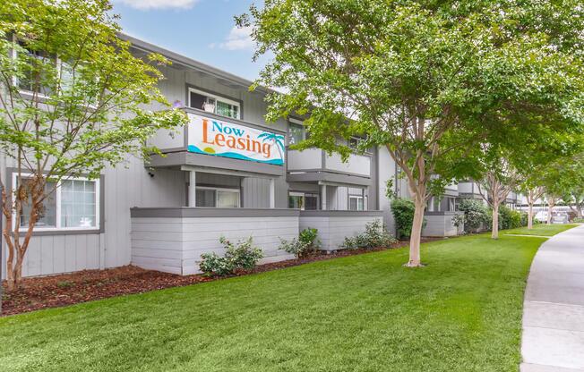 A row of apartment units with a "Now Leasing" sign prominently displayed. The building has light gray siding and is surrounded by manicured green lawns and landscaping. Trees and shrubs add a natural touch to the scene, which features a clear blue sky above.