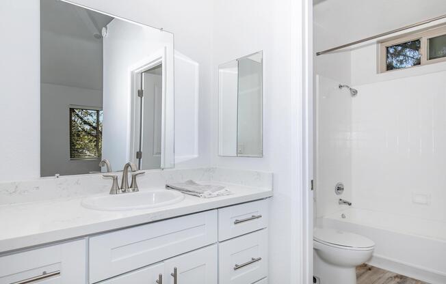 Modern bathroom with a dual sink vanity featuring a white countertop and ample storage. A large mirror above the sinks reflects the bright, airy space. The bathroom includes a shower stall with white walls and a toilet, set against light wood-style flooring. Natural light filters in through a window.