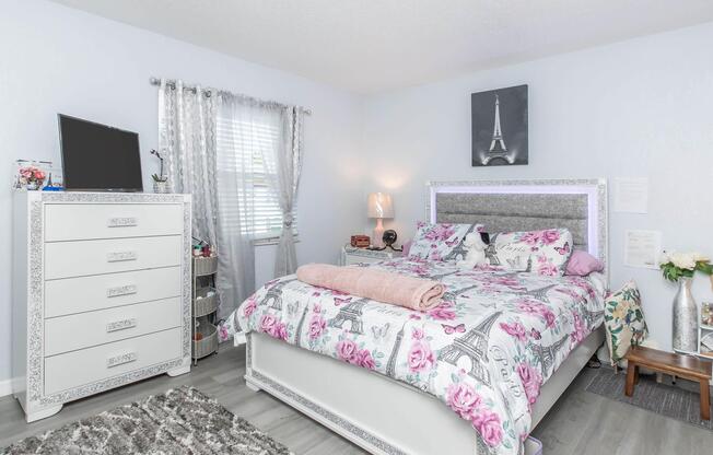 A cozy bedroom featuring a floral-patterned bedspread on a bed with a pink blanket. There's a white dresser with ornate details, a television on a stand, and light-colored curtains. A small vase of flowers and a picture of the Eiffel Tower add charm to the decor. The room has a light and airy feel.
