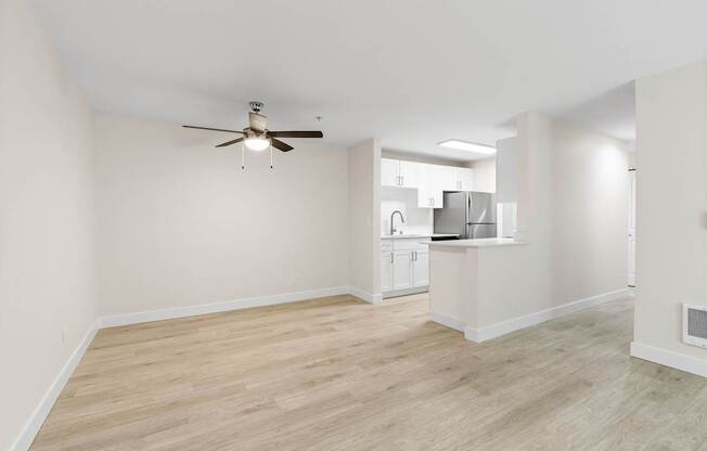 A room with a ceiling fan and light fixture, and a kitchen area in the background.