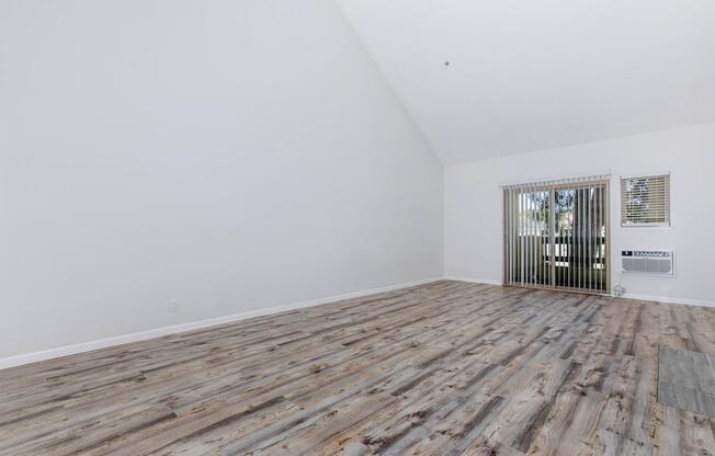 A spacious, well-lit room with high ceilings and wooden flooring. There are large windows with vertical blinds revealing outdoor greenery. An air conditioning unit is visible on the wall, and the room is mostly bare, offering a blank canvas for decorating.