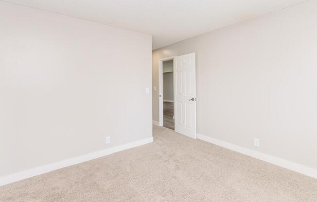 A sparse room with light beige carpet and plain white walls. There's an open door leading to another room, and the space is well-lit, creating a clean and neutral ambiance. No furniture is present, giving it an open and airy feel.