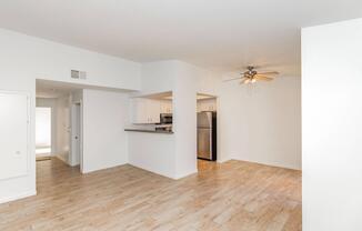 Spacious living area with light wood flooring, ceiling fan, and modern appliances. Includes a galley-style kitchen visible in the background, along with a doorway leading to another room. Walls are painted white, creating a bright and airy atmosphere.