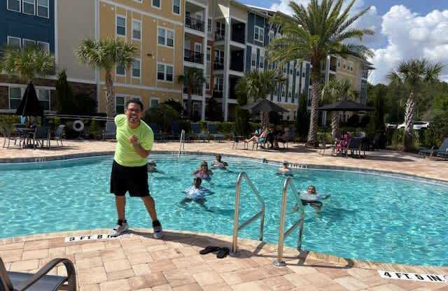 A man in a yellow shirt is standing by a pool with people swimming.