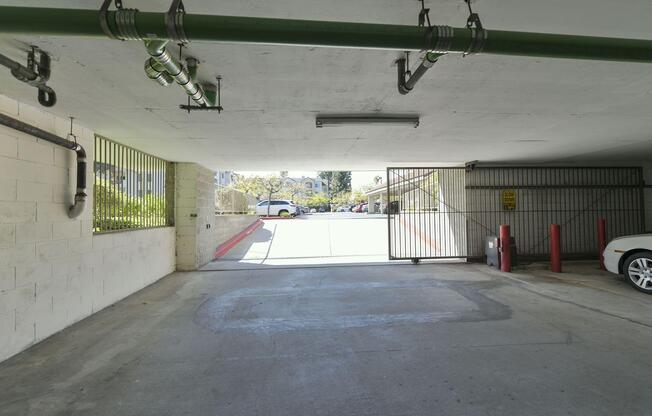 A view from inside an underground parking garage, showing an exit with a gated entrance. Green pipes run along the ceiling, and there are red poles marking the area. Outside, a paved area with parked cars and greenery can be seen in the distance. Natural light illuminates the scene.