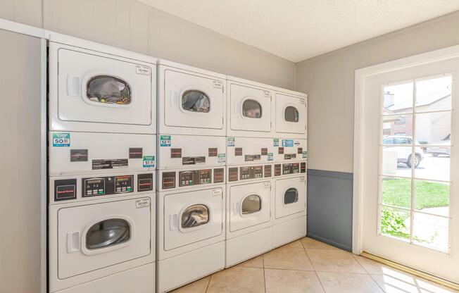 A laundry room featuring a row of white commercial washers and dryers against a light-colored wall. To the right, there is a window with several panes, allowing natural light to brighten the space. The floor is tiled, and there is a glimpse of greenery outside through the window.