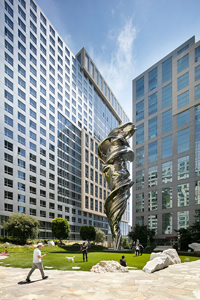 a large metal sculpture in a park in front of two large buildings