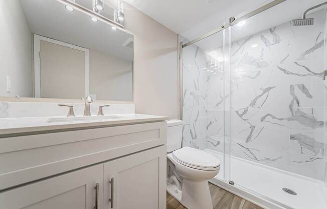 A white toilet sits in a bathroom with a marble shower wall.