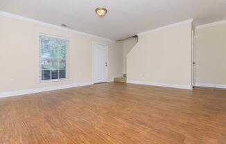 Spacious, empty living room with light-colored walls and a large window letting in natural light. The floor is wooden and there is a ceiling light fixture. A closed door is visible, along with a staircase leading to a second level, suggesting an inviting and well-maintained interior.