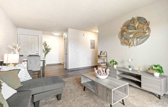 A living room with a grey couch, a coffee table, and a large sculpture on the wall A row of apartment buildings with green lawns in front A bedroom with a bed, nightstand, and window at Wildflower, Kennewick, WA 99336..