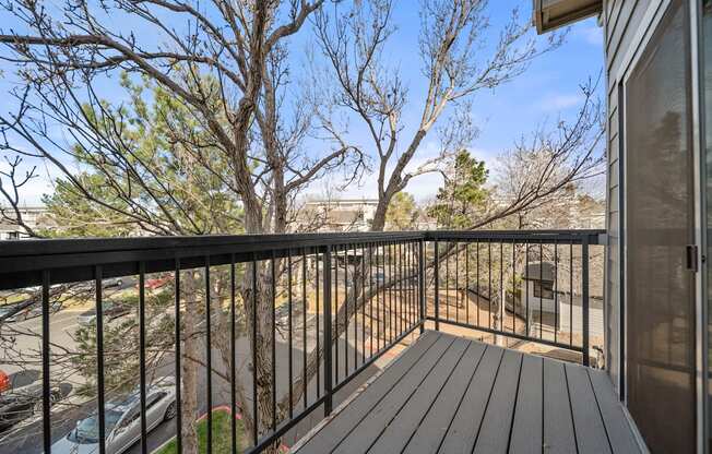 A balcony with a black railing and a wooden floor overlooks a parking lot and trees.