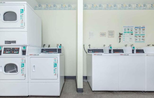 A clean, well-lit laundry room featuring stacked washing machines and dryers on the left, and row of front-loading washers with coin slots on the right. The walls are painted a light color with a decorative border, and there are informative signs and instructions displayed nearby.