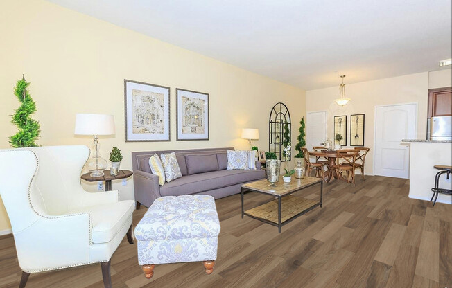 A bright and modern living room featuring a gray sofa with decorative pillows, a light-colored armchair, and a patterned ottoman. There is a glass coffee table on a wooden floor, framed artwork on the wall, and a dining area in the background with a round table and chairs. Green plants add a touch of nature.